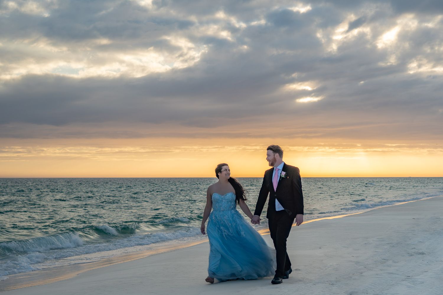 Elegant couple holding hands and walking along a white sand beach at sunset in formal wedding attire.