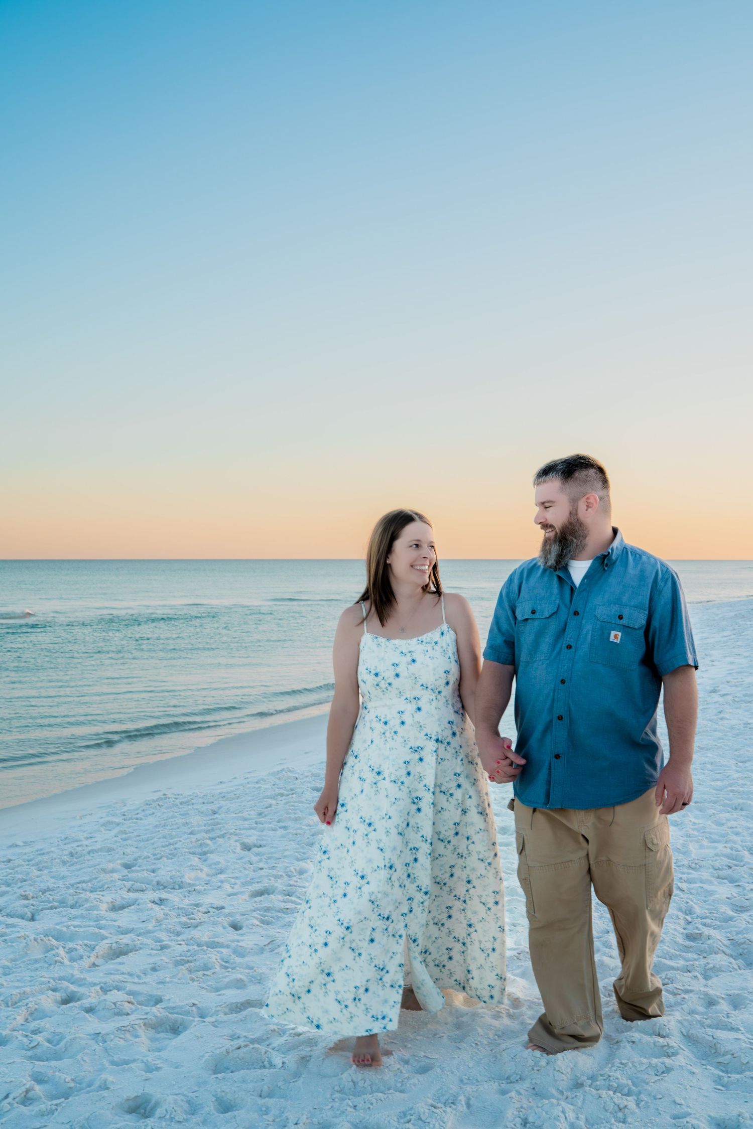 Couple holding hands and walking on a white sand beach at sunset, woman in floral dress, man in teal shirt.