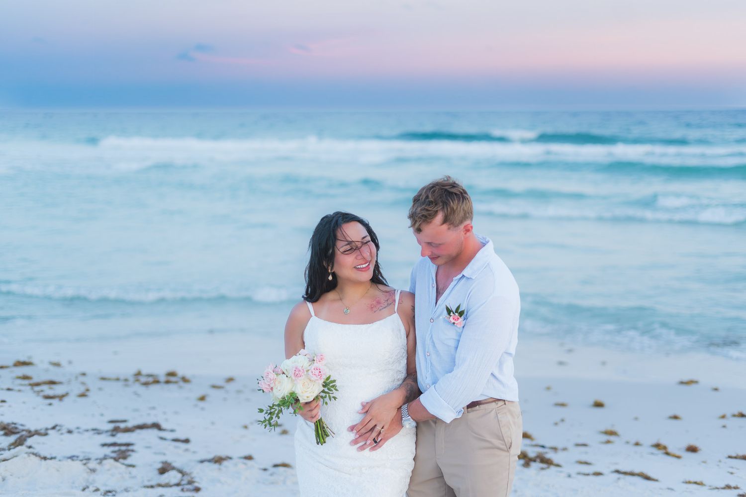 Bride and groom share a romantic beach wedding moment at sunset, with ocean waves and pastel sky in the background.