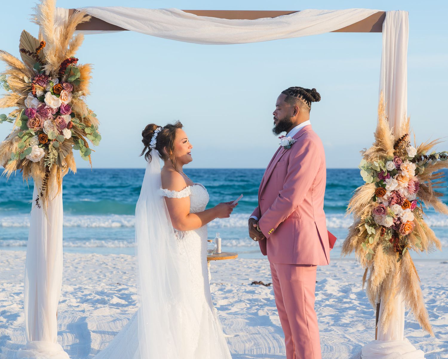 Bride and groom share a romantic moment under a boho floral arch during a sunny beach wedding ceremony.