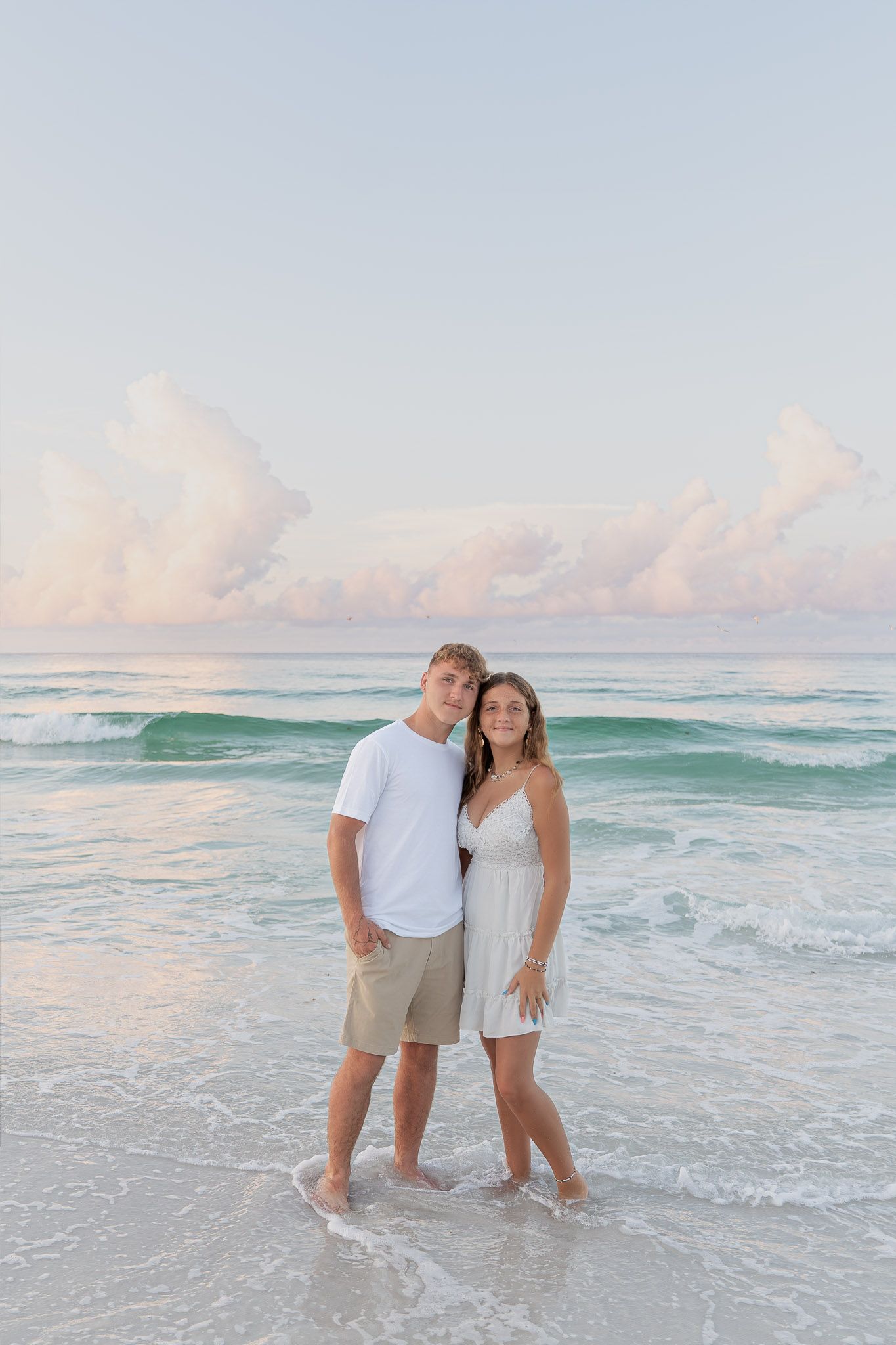 Young couple standing barefoot in ocean waves on a sandy beach at sunset, wearing white outfits.