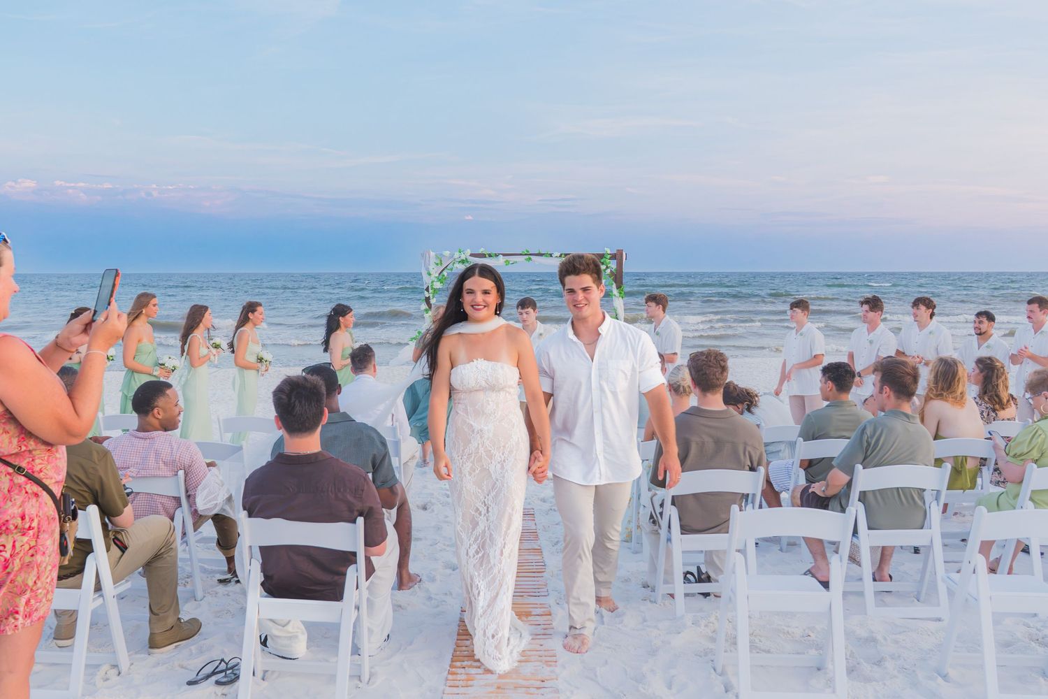 Couple walks hand-in-hand after beach wedding ceremony with guests seated on white chairs at sunset.