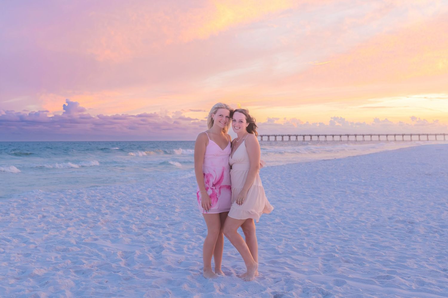 Two women embrace on a white sand beach at sunset with pink and purple skies.