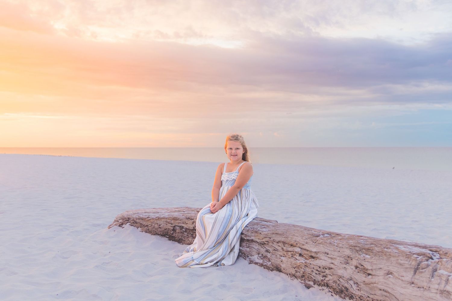 Young blonde woman in a flowing white dress sitting on driftwood at a serene white sand beach during a pastel sunset.