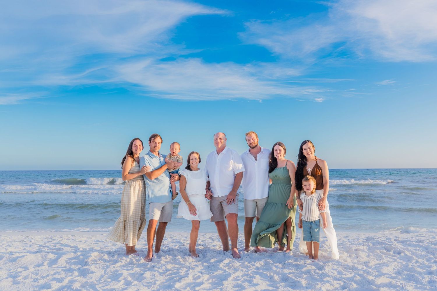 Family portrait on white sand beach with blue sky, adults and children posing together near ocean waves.