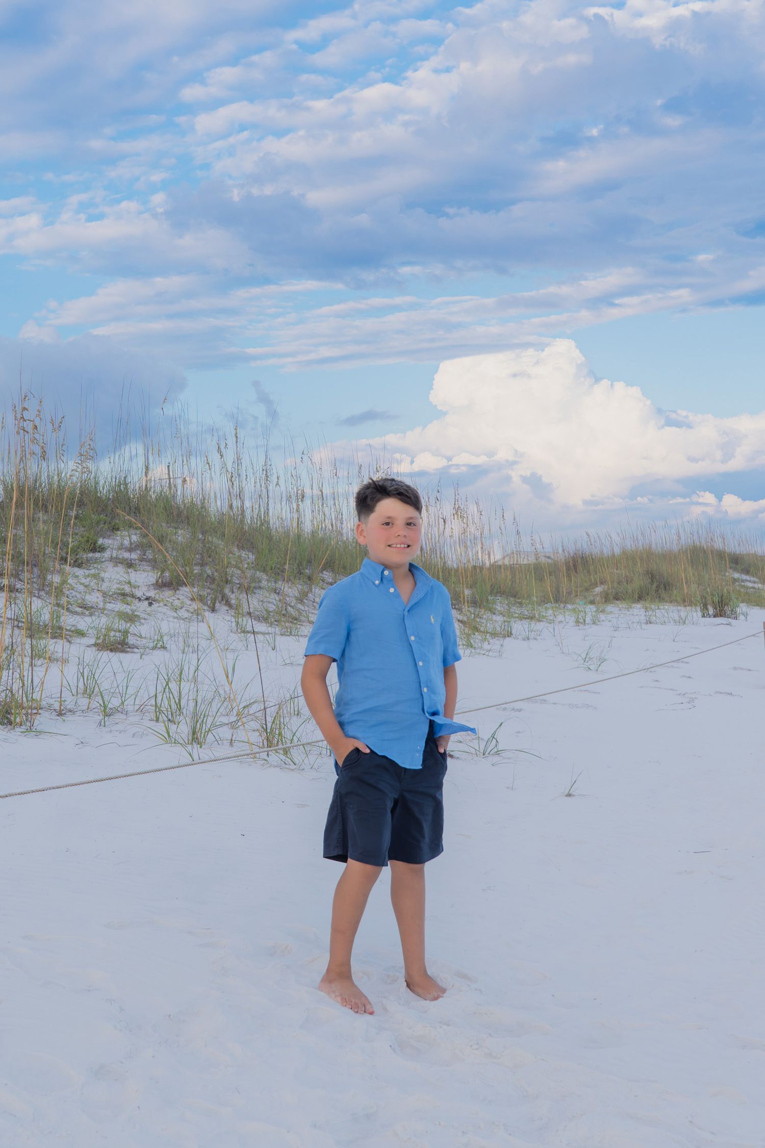 A smiling boy in a blue shirt and navy shorts stands barefoot on white sand dunes with sea oats and clouds behind him.