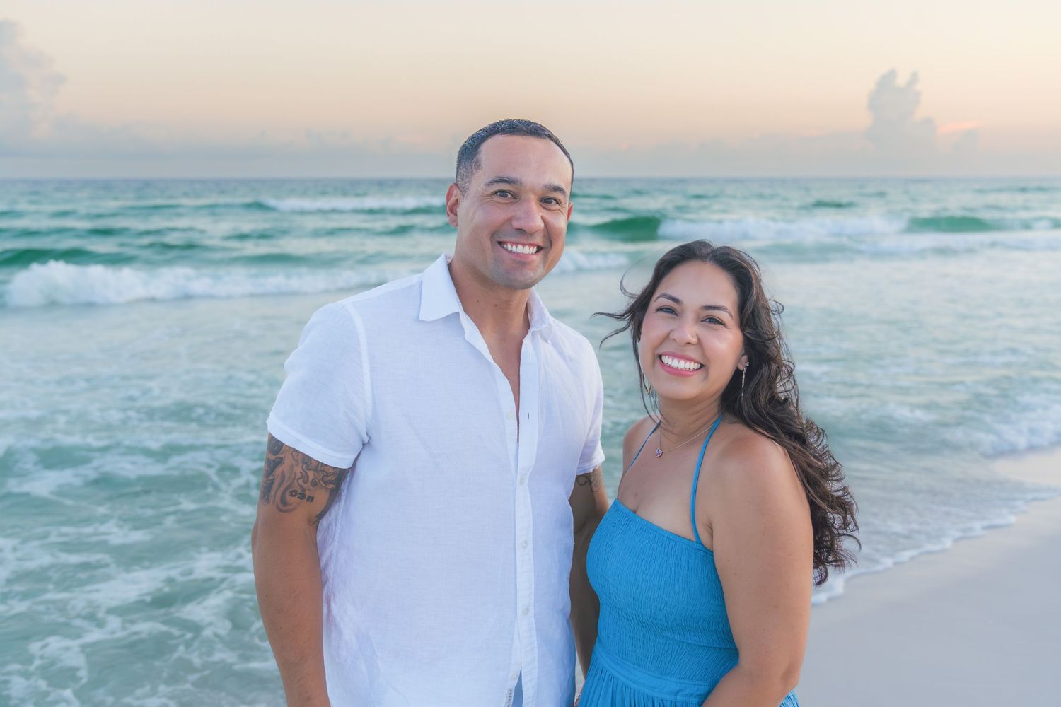 Smiling couple posing together on a tropical beach at sunset with turquoise ocean waves.