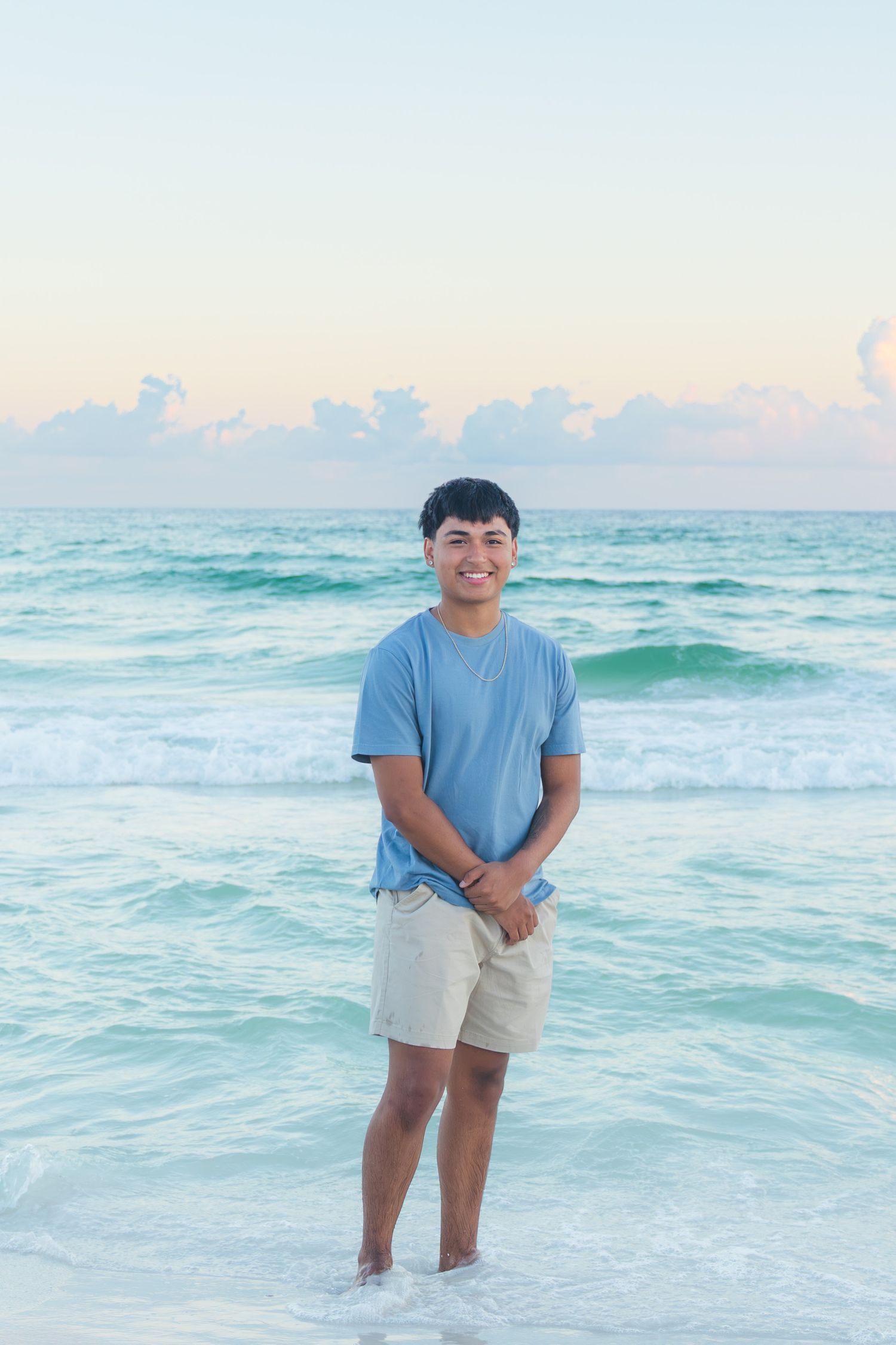 Smiling young man standing in ocean waves at sunset wearing a blue t-shirt and khaki shorts at a tropical beach.