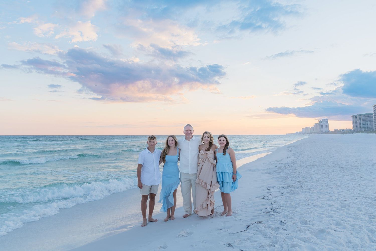 Family of five poses on a white sand beach at sunset with pastel skies and turquoise Gulf waters behind them.