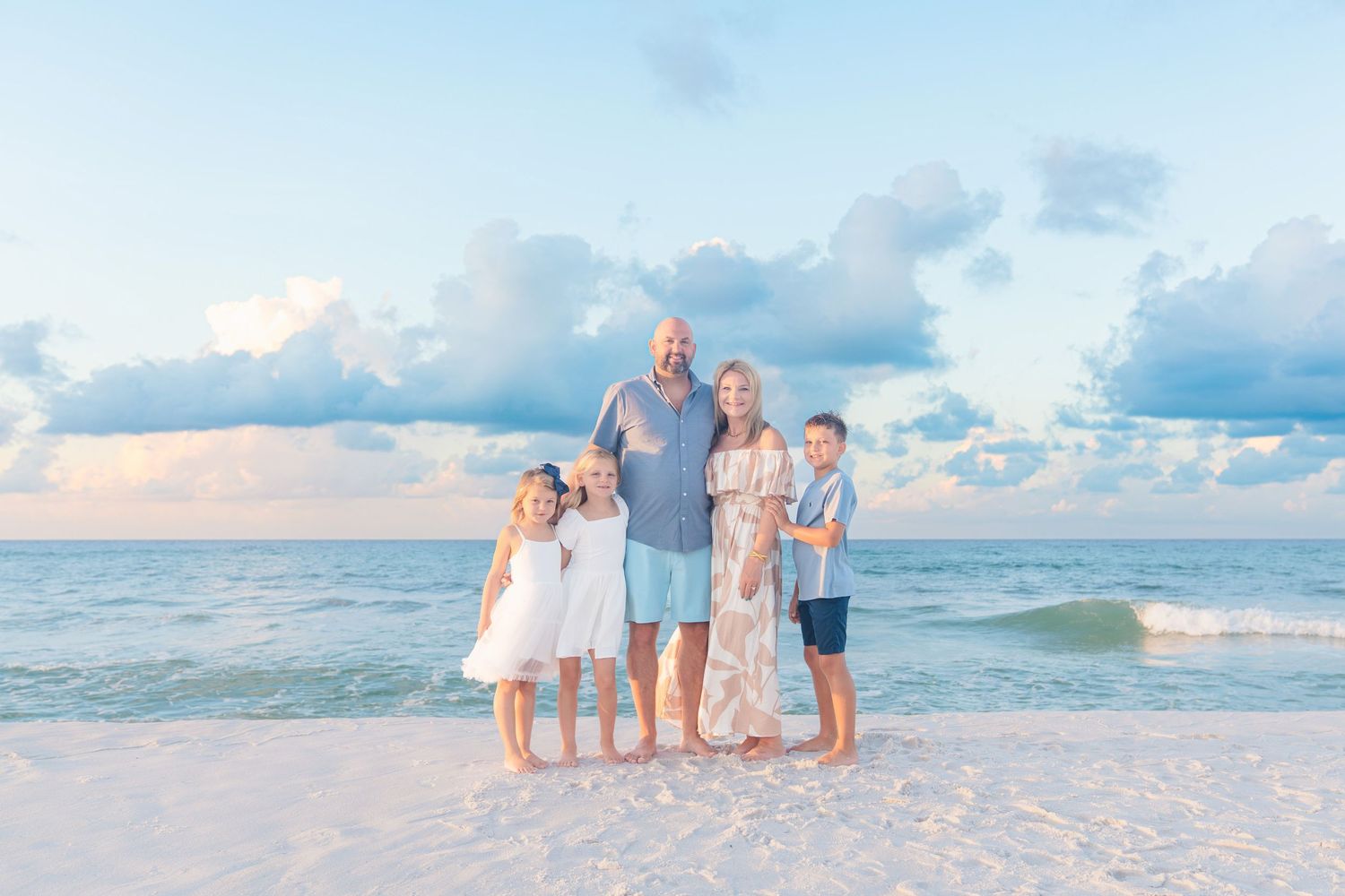 Family of five posing on a white sand beach at sunset with calm turquoise ocean waves and pastel cloudy sky.