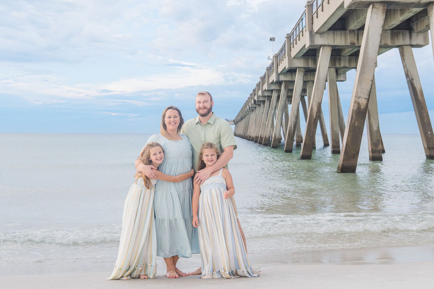 Family of four poses on sandy beach near pier, wearing coordinating pastel outfits, with calm ocean waves in background.