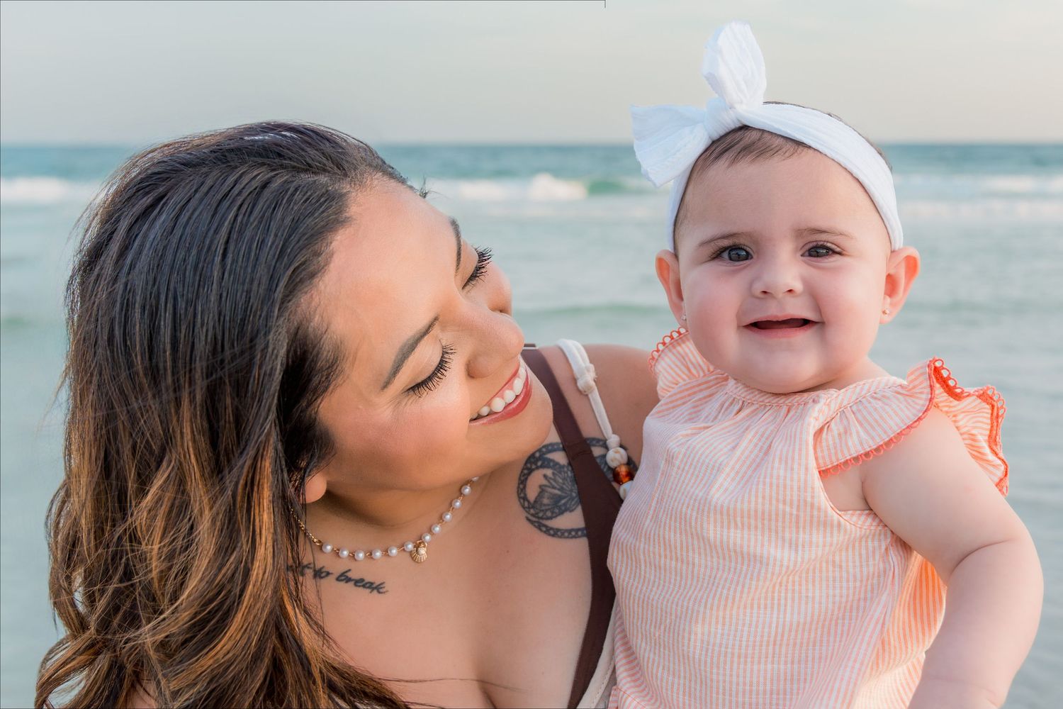 A mother holds her smiling baby girl in a pink dress and white bow headband at the beach.