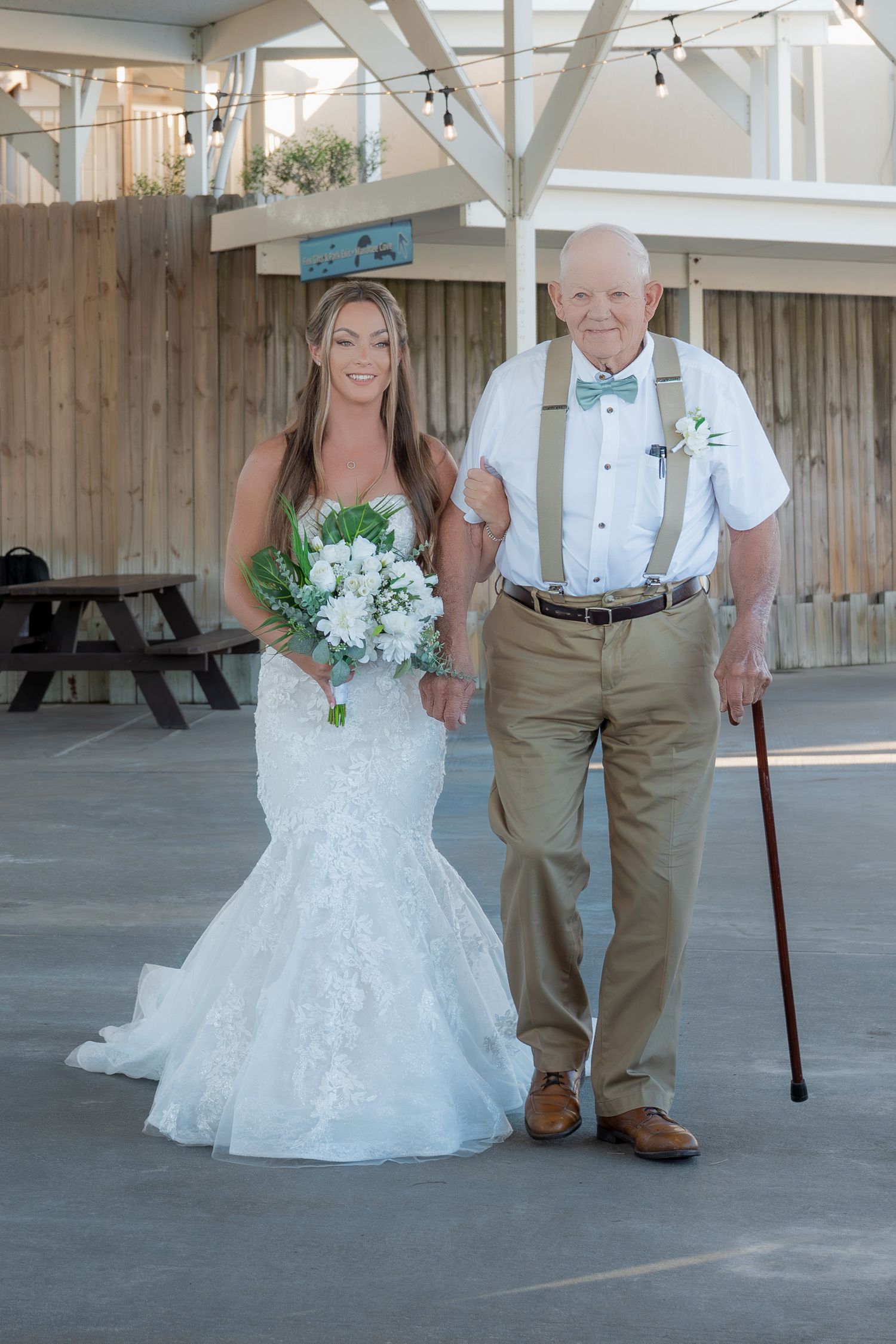 A smiling bride in a mermaid gown holds white flowers beside an elderly man with suspenders and a cane.