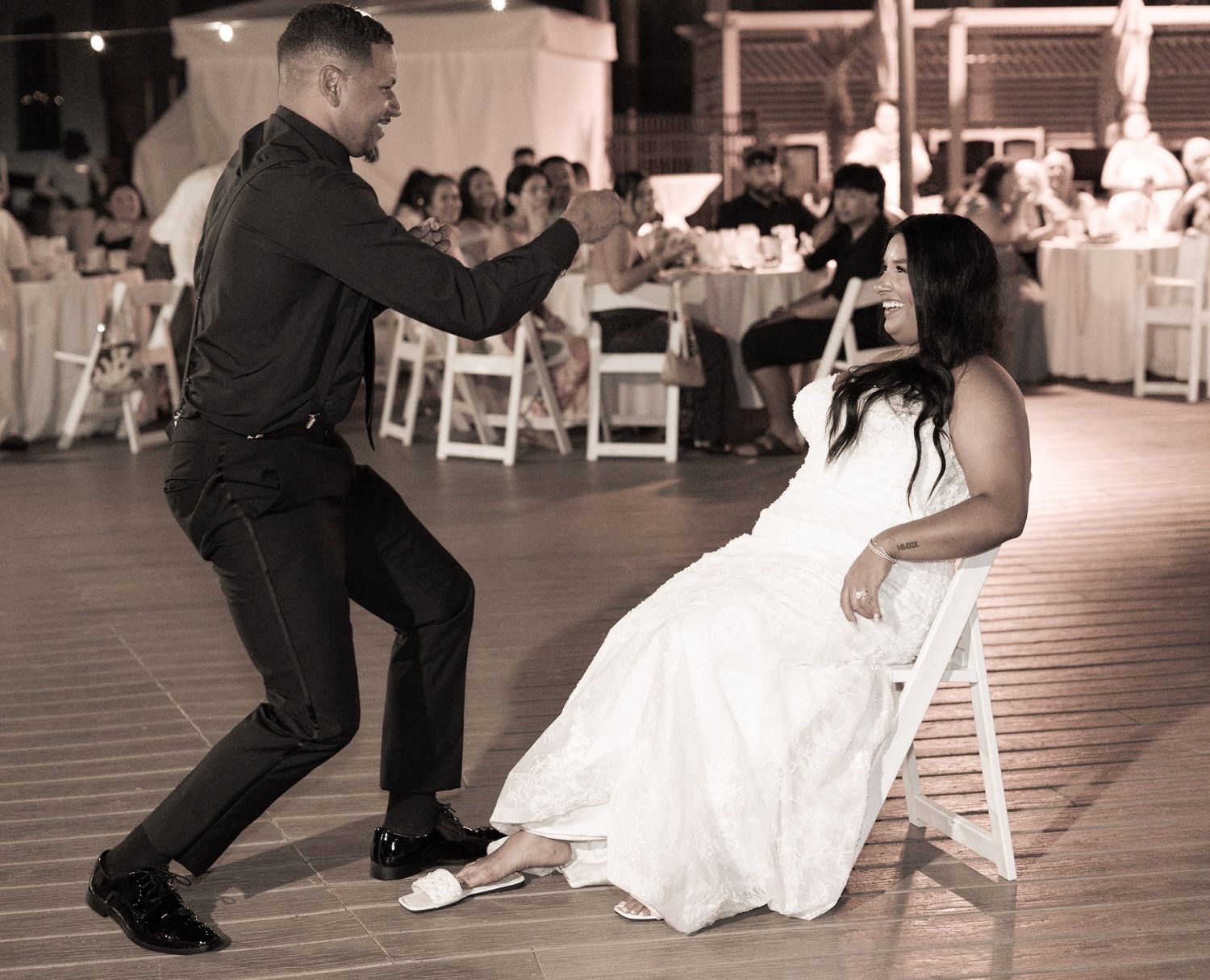 Groom performs a lively garter removal dance for the seated bride at the wedding reception in sepia tone.