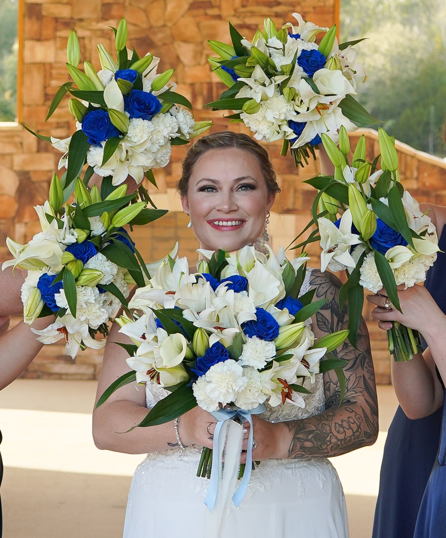 Smiling bride surrounded by bridesmaids holding blue and white floral bouquets at an outdoor wedding ceremony.