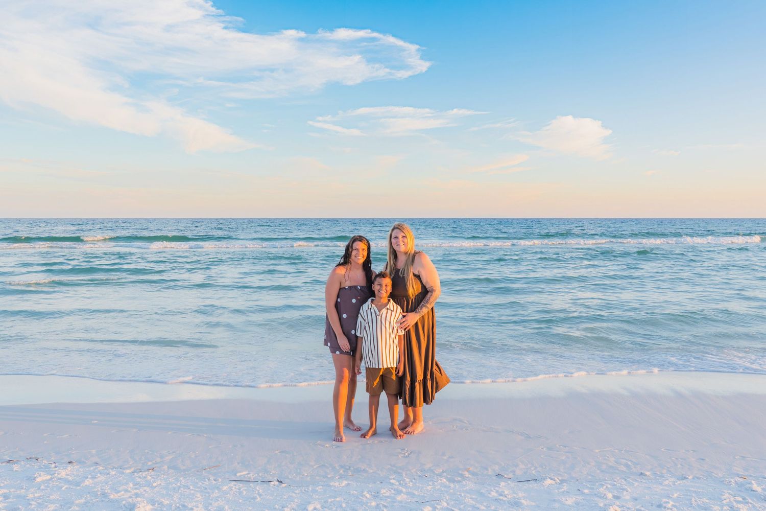 Family of three posing on a white sand beach at sunset with calm turquoise ocean waters in the background.