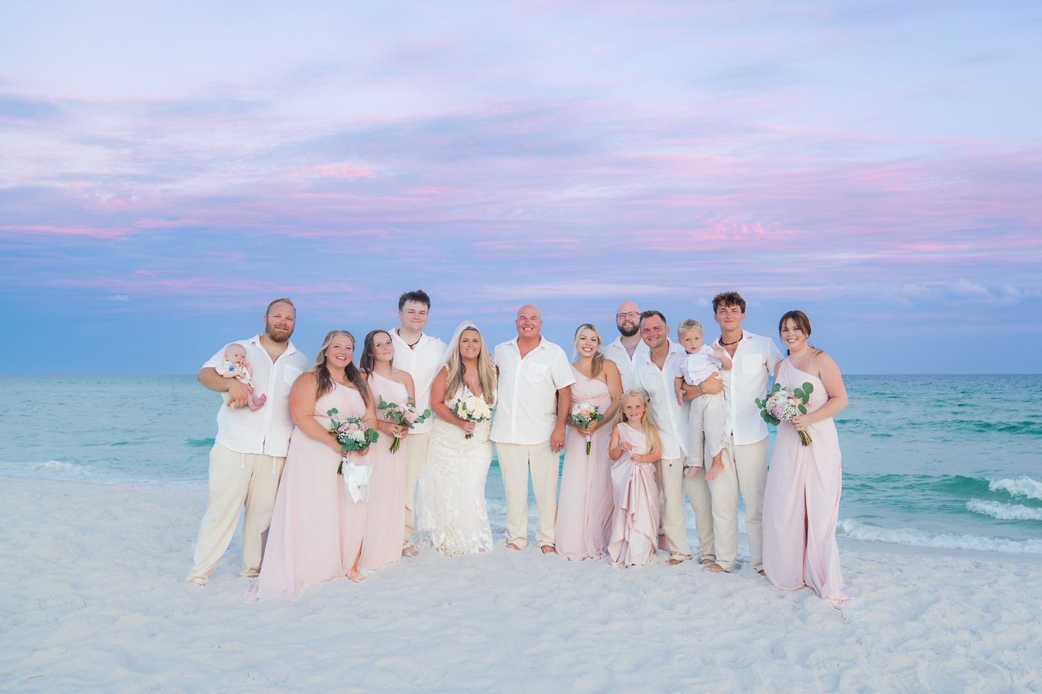 Wedding party poses on a white sand beach at sunset with pink and purple sky, bride in white, attendants in blush gowns.