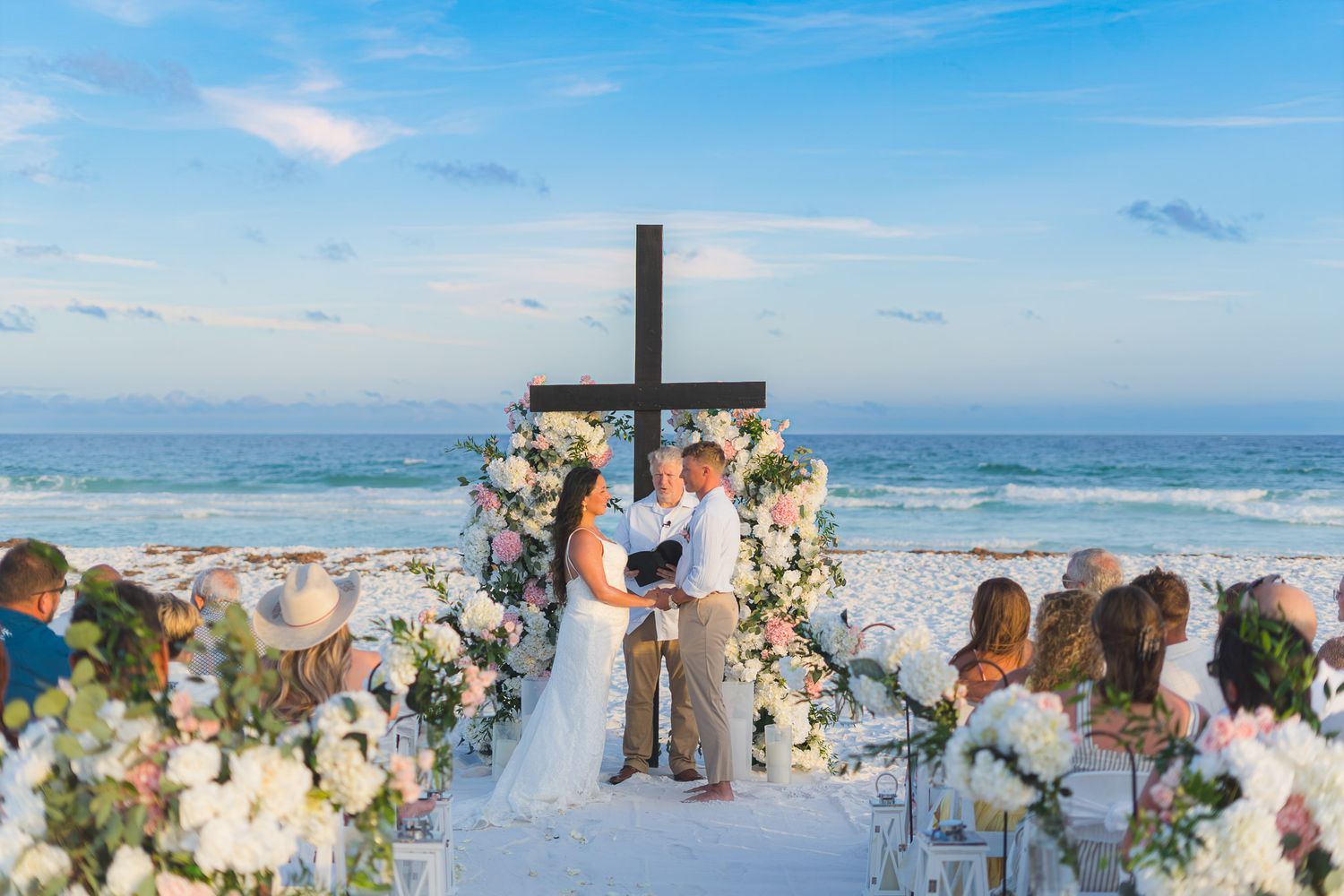 Romantic beach wedding ceremony with floral arch and cross at ocean sunset.
