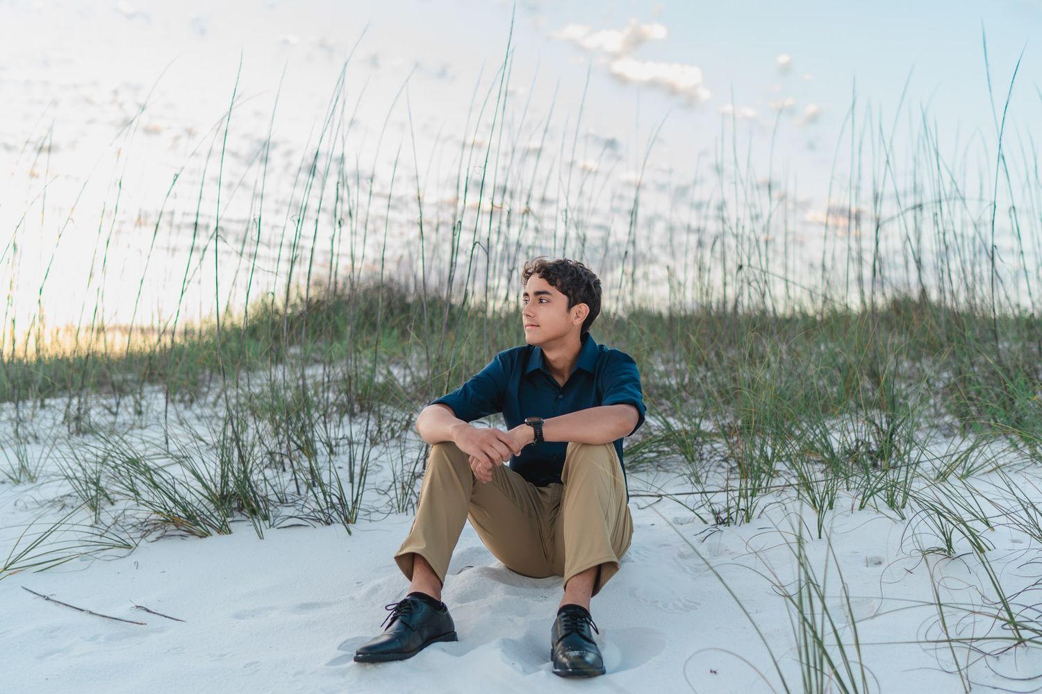 Young man in navy shirt and khaki pants sitting on white sand dunes with sea oats at a serene beach.