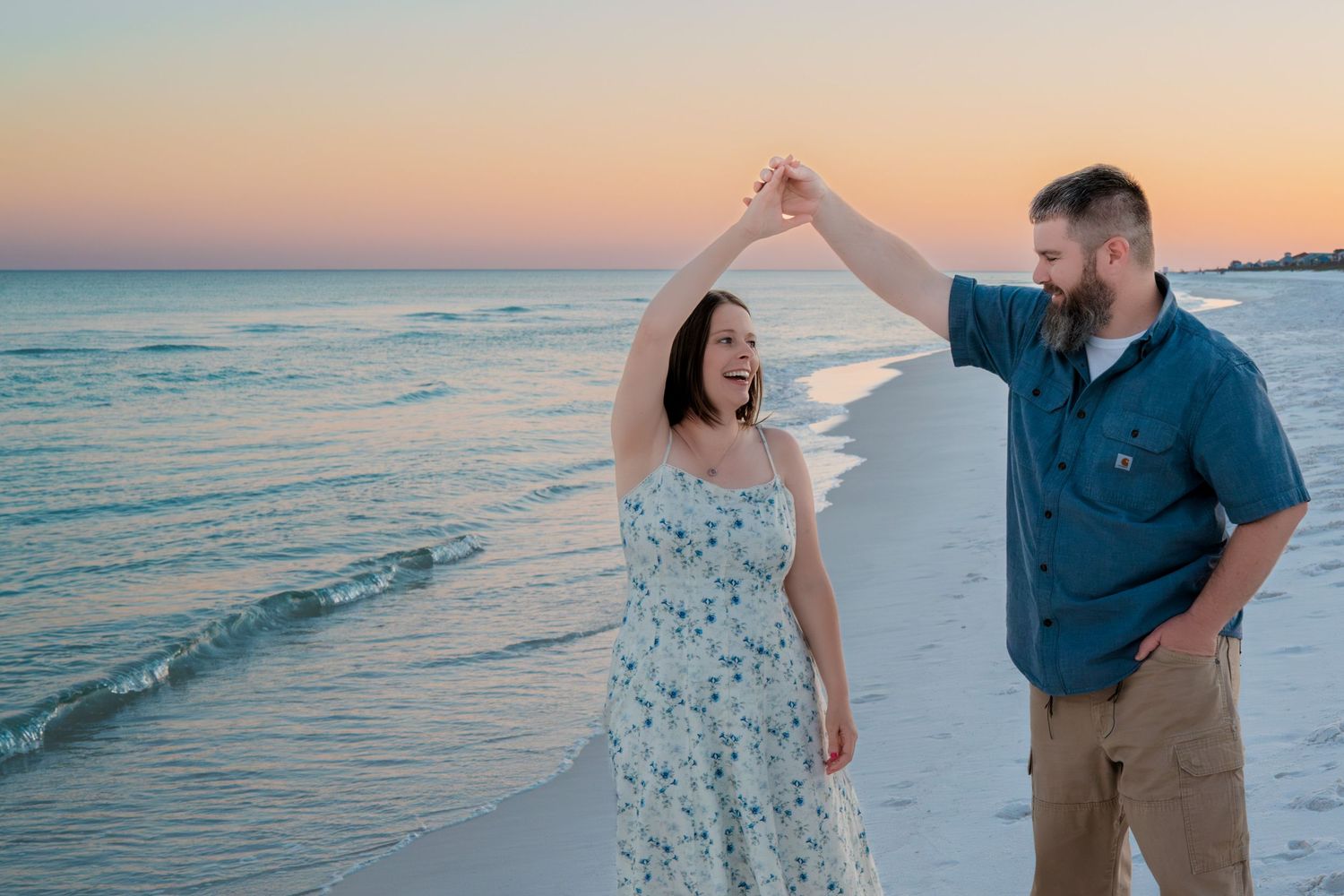 Couple dancing on beach at sunset, man spinning woman in floral dress near ocean shoreline.