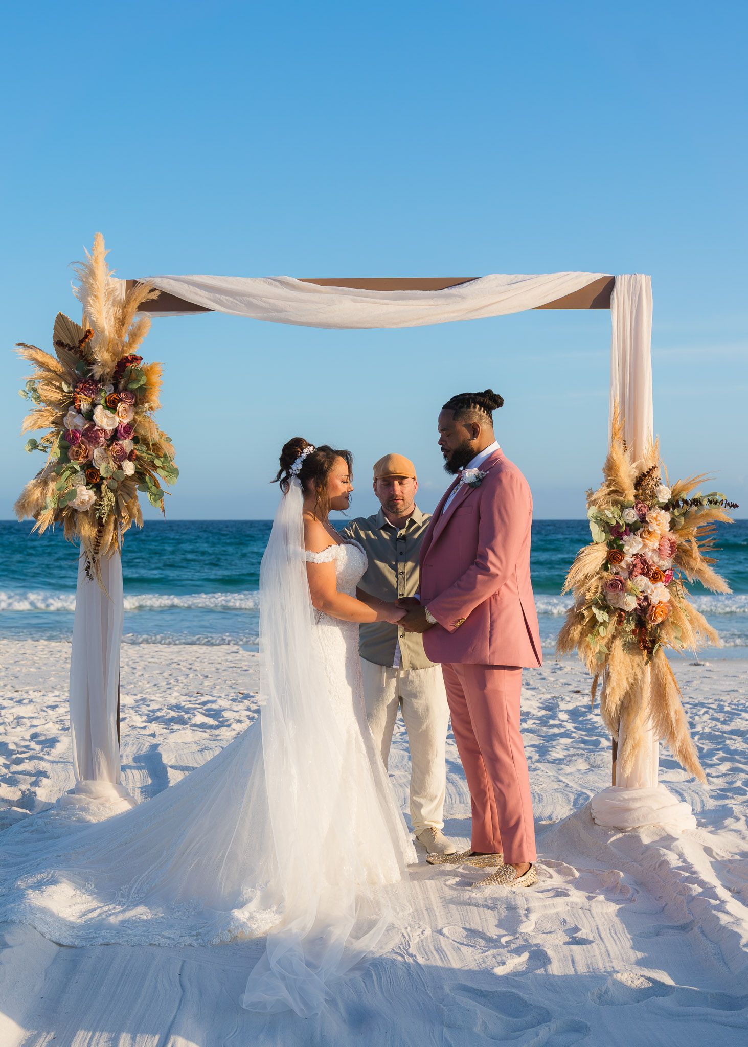 Couple exchanging vows at a beach wedding under a decorated arch with floral arrangements, ocean backdrop.