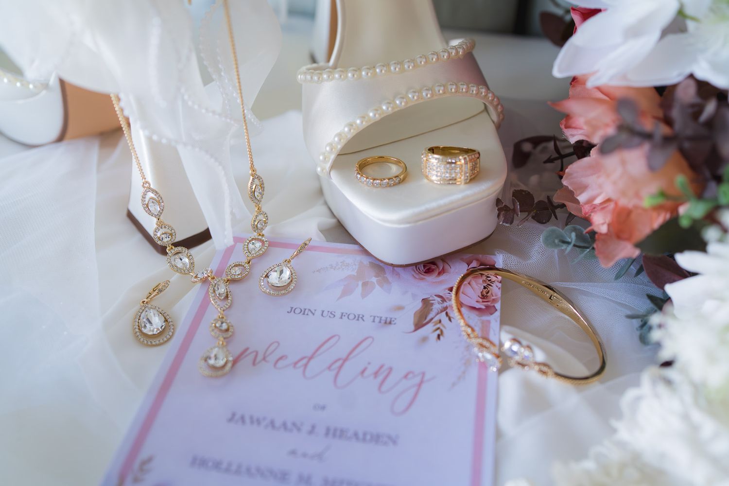 Gold wedding jewelry set with necklace, bracelet, rings in velvet box, heels, and floral invitation on white background.