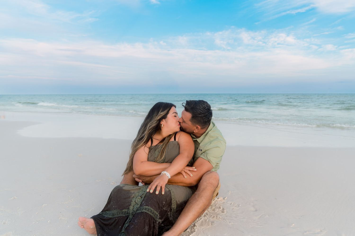 Couple sharing a romantic kiss while sitting on a white sandy beach with calm turquoise ocean waves behind them.
