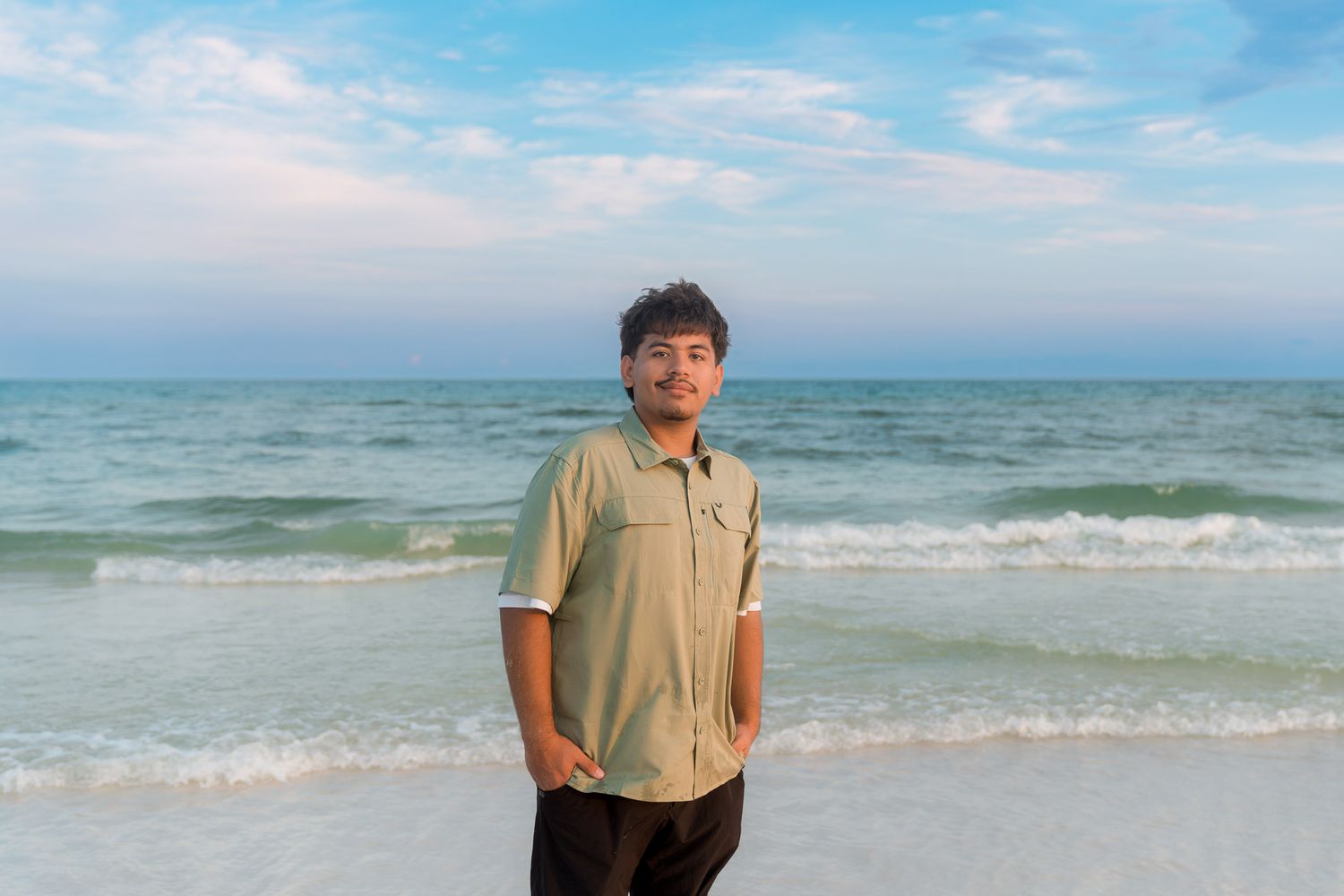 Young man smiling on a sandy beach with turquoise ocean waves and a pastel blue sky behind him.