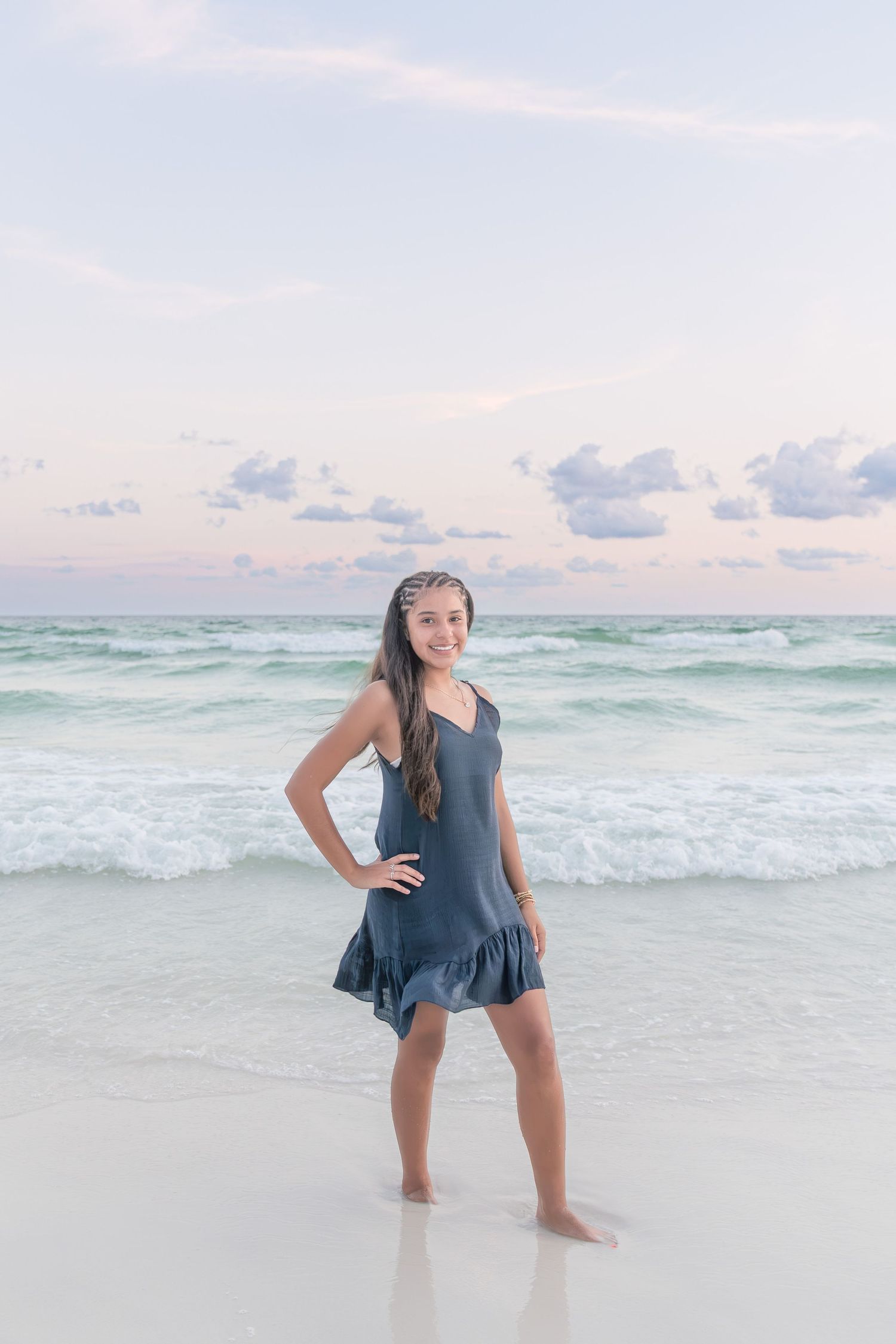 Young girl in gray ruffle dress stands barefoot on sandy beach with turquoise ocean waves and pastel sky behind her.