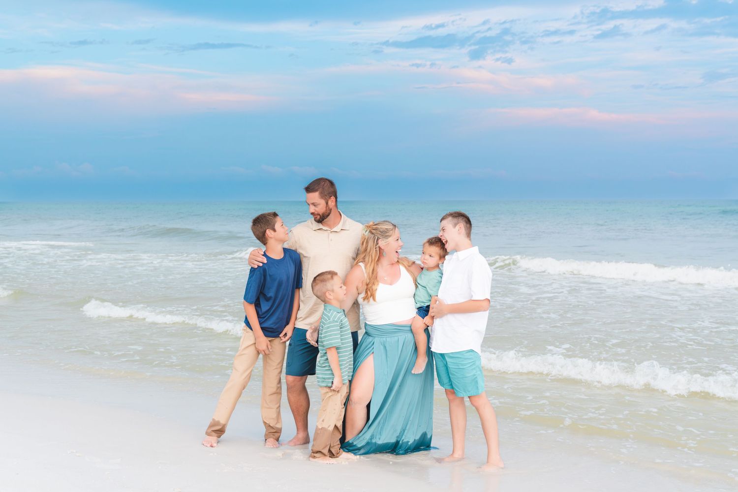 Family of six posing together on a sandy beach at sunset with gentle ocean waves in the background.