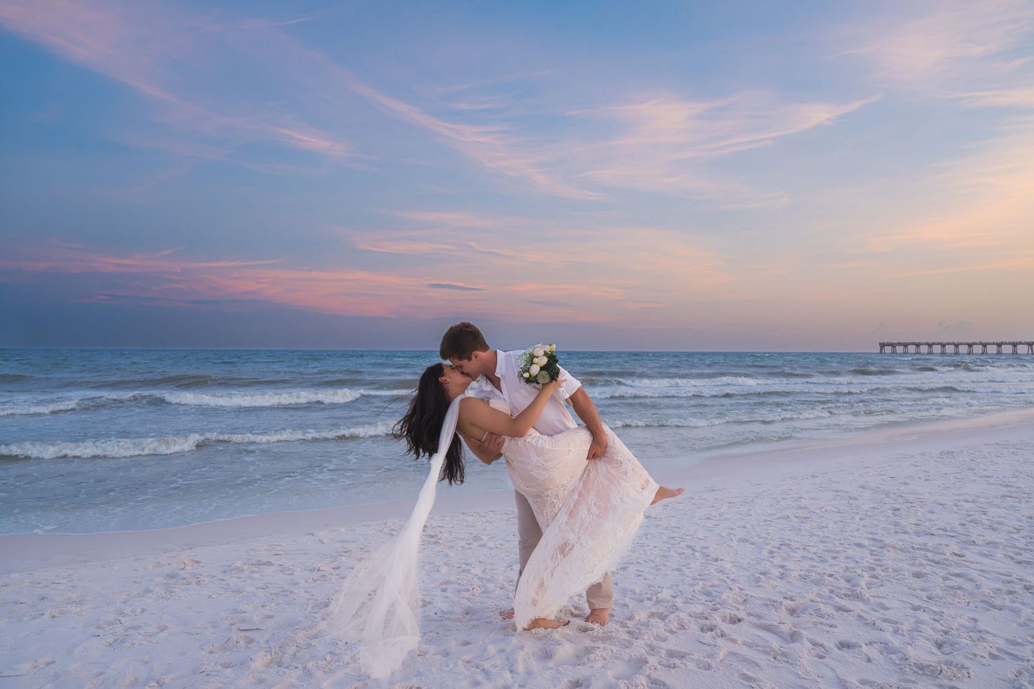 Newlywed couple sharing a romantic kiss on a white sand beach at sunset with a pier in the background.