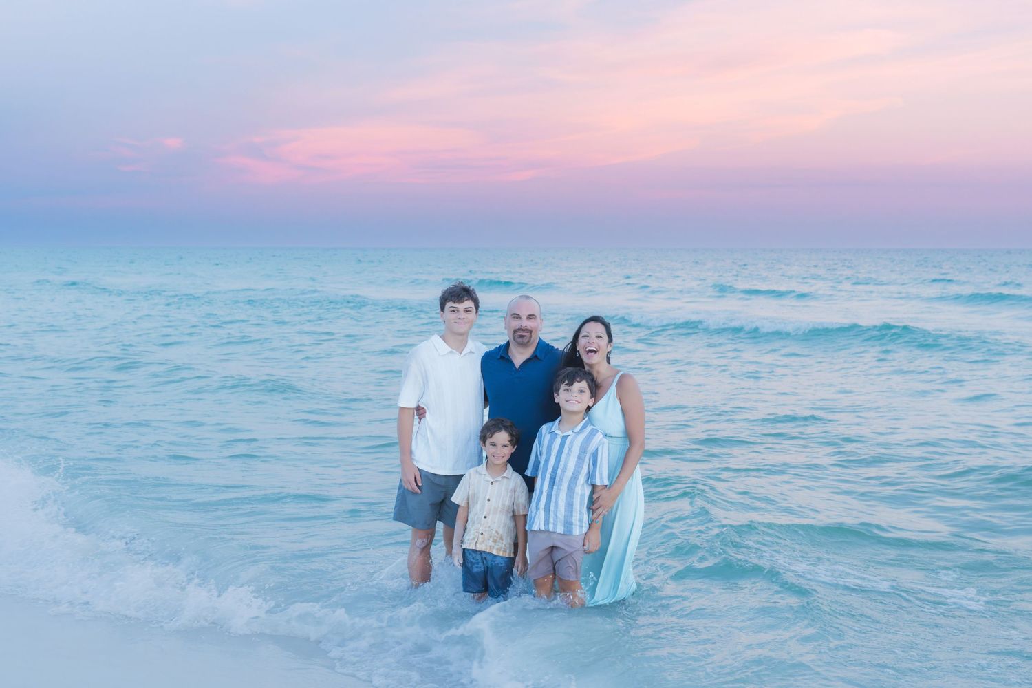 Family of five posing together in shallow ocean waves at sunset with pink and blue sky backdrop.