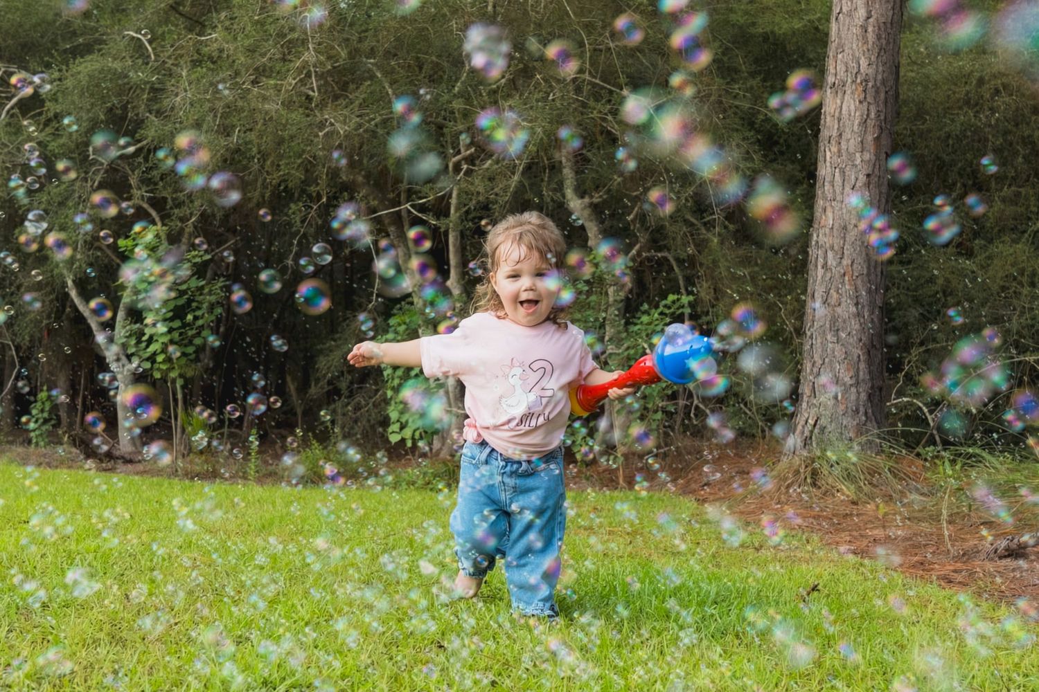 A happy toddler girl plays outdoors with a red bubble gun surrounded by floating colorful bubbles.