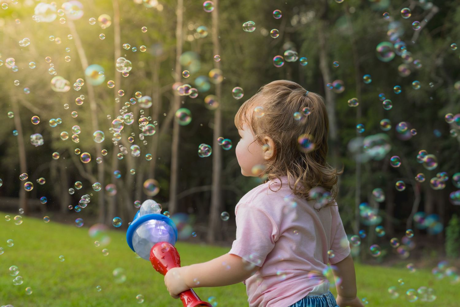 A young girl uses a bubble gun toy outdoors, surrounded by floating soap bubbles in a sunny green park.