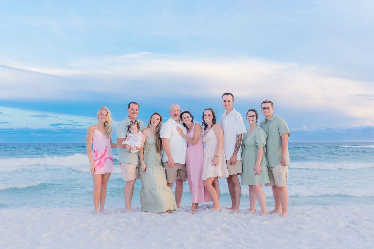 Large family group posing together on a white sand beach at sunset wearing pastel outfits with ocean waves behind them.