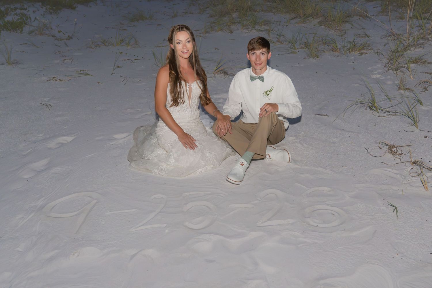 Bride and groom sitting on white sand beach with wedding date written in sand at night.