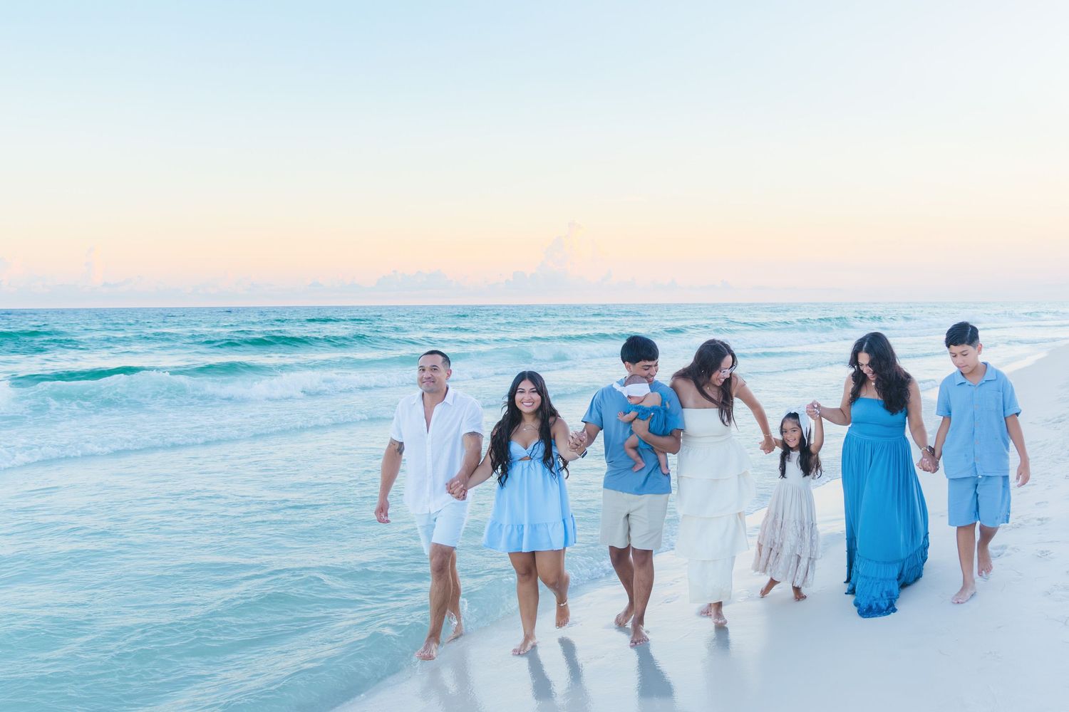 Family walking hand-in-hand on a white sand beach at sunset, wearing white and blue outfits with ocean waves behind them.