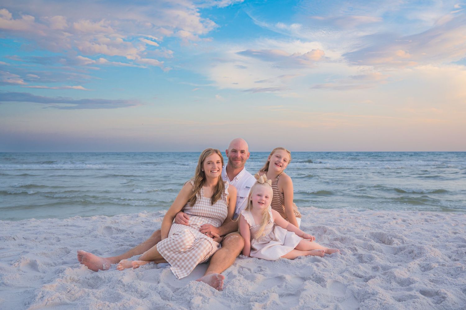 Family of four posing together on white sand beach at sunset with calm ocean waves in the background.
