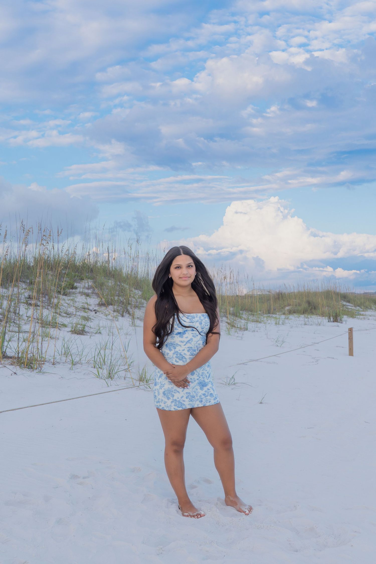 Young woman in blue floral mini dress posing barefoot on white sand beach with sea oats and cloudy sky.