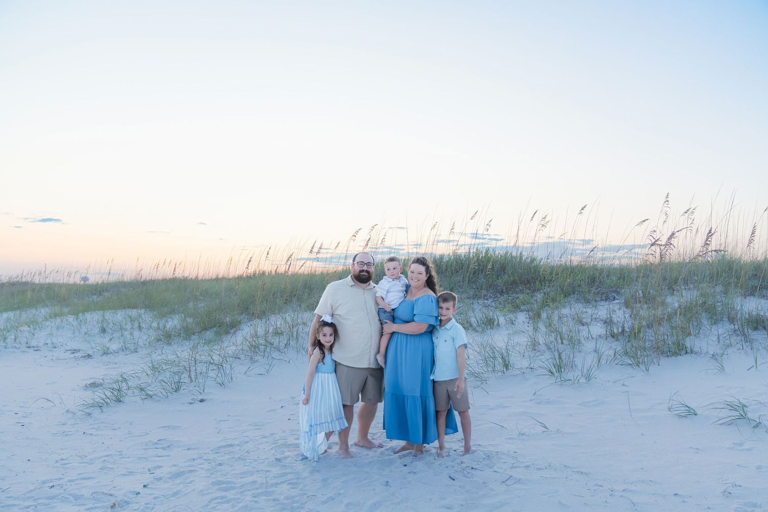 Family of five posing on sand dunes with sea oats at golden hour on a beautiful beach.