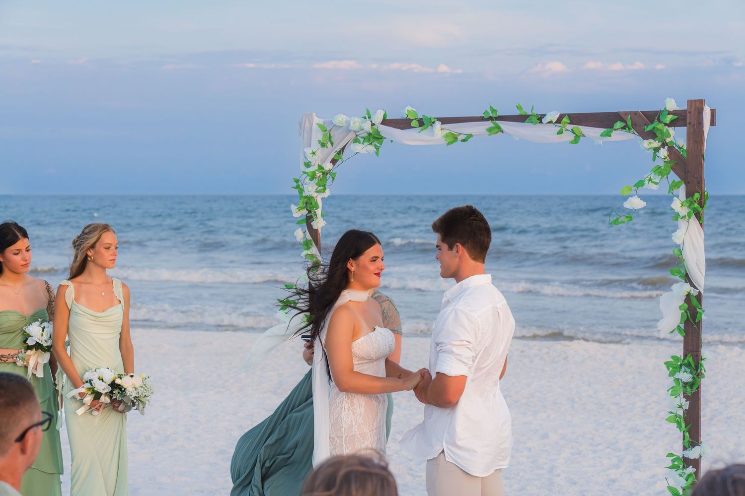 Couple exchanges vows at a romantic beach wedding ceremony under a floral arch with bridesmaids nearby.