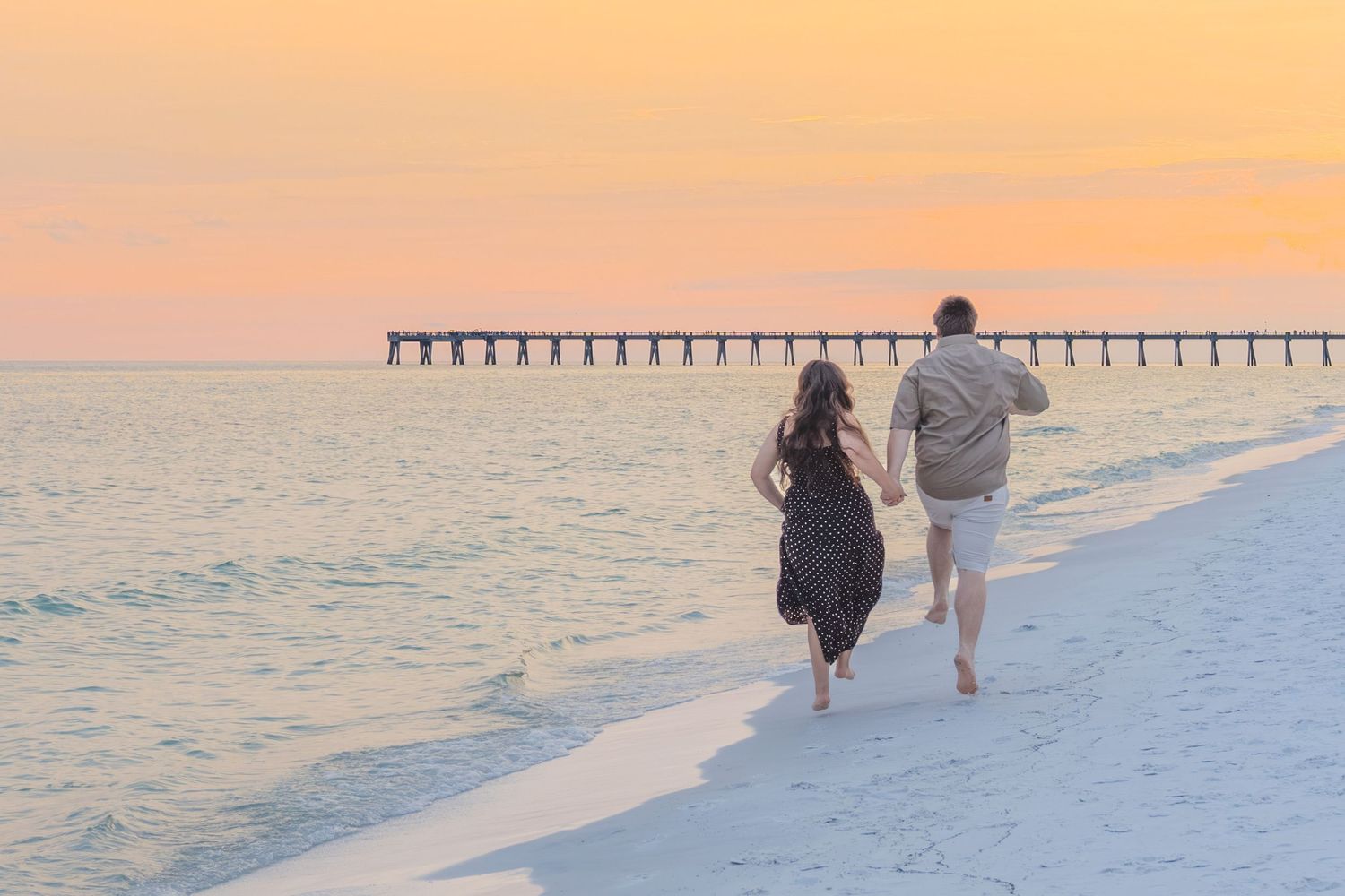 Couple holding hands running along a white sand beach at golden sunset with a pier in the distance.
