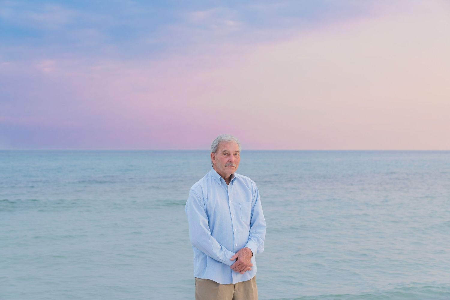 Elderly man standing on a beach at sunset wearing a light blue shirt with pastel sky and ocean backdrop.