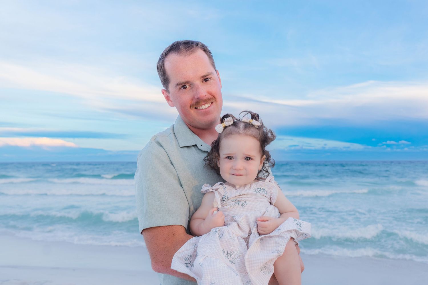 A smiling man holds a young girl in a white dress on a sandy beach at sunset.