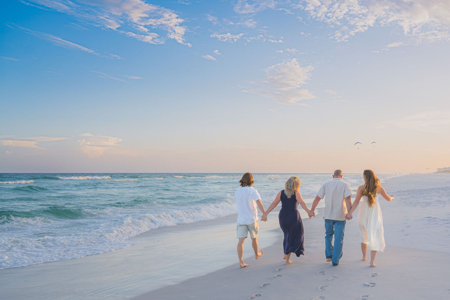 Family of four holding hands walking along a sandy beach at sunset with turquoise waves.