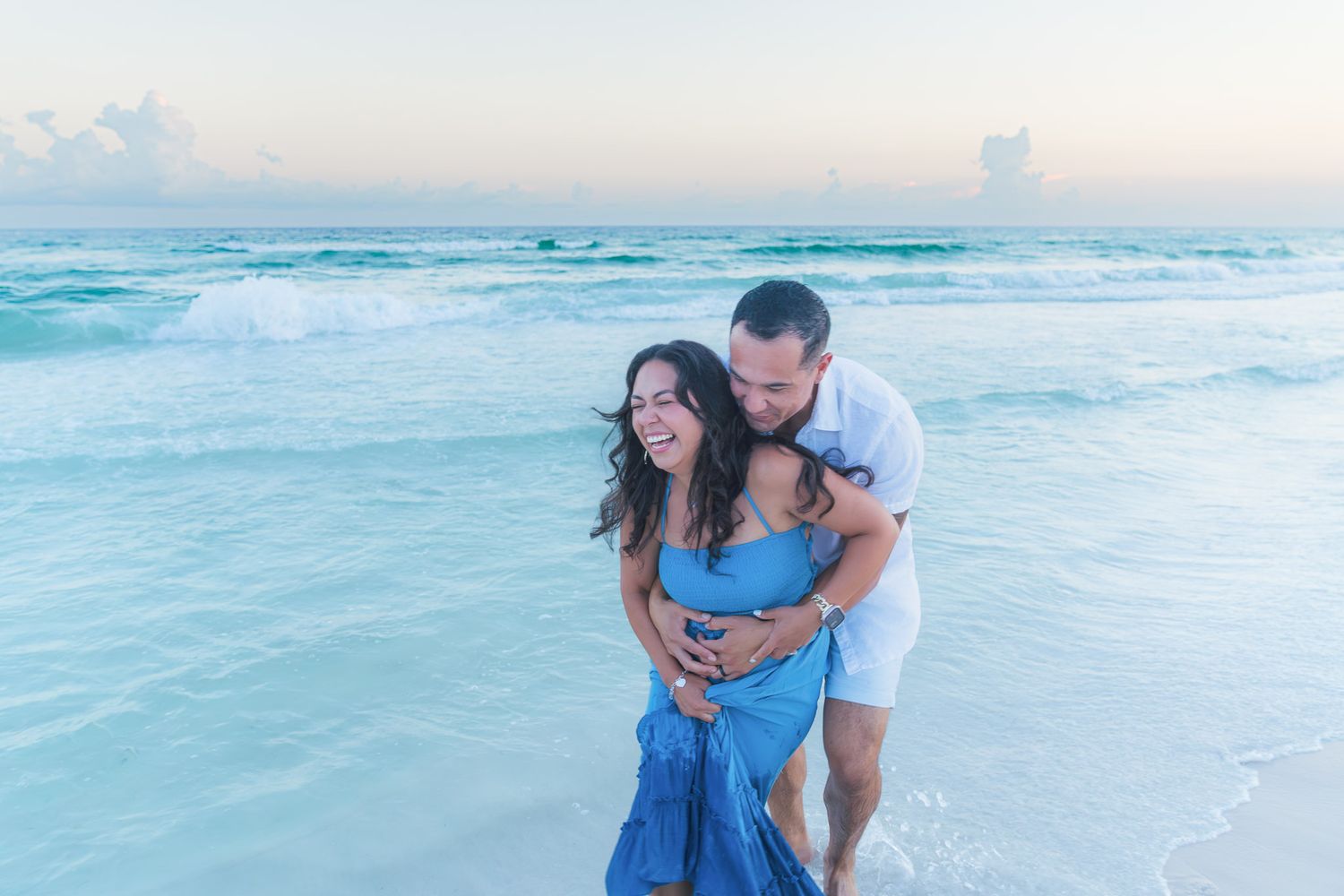 Couple embracing and laughing on a tropical beach at sunset, woman in blue dress, waves in background.