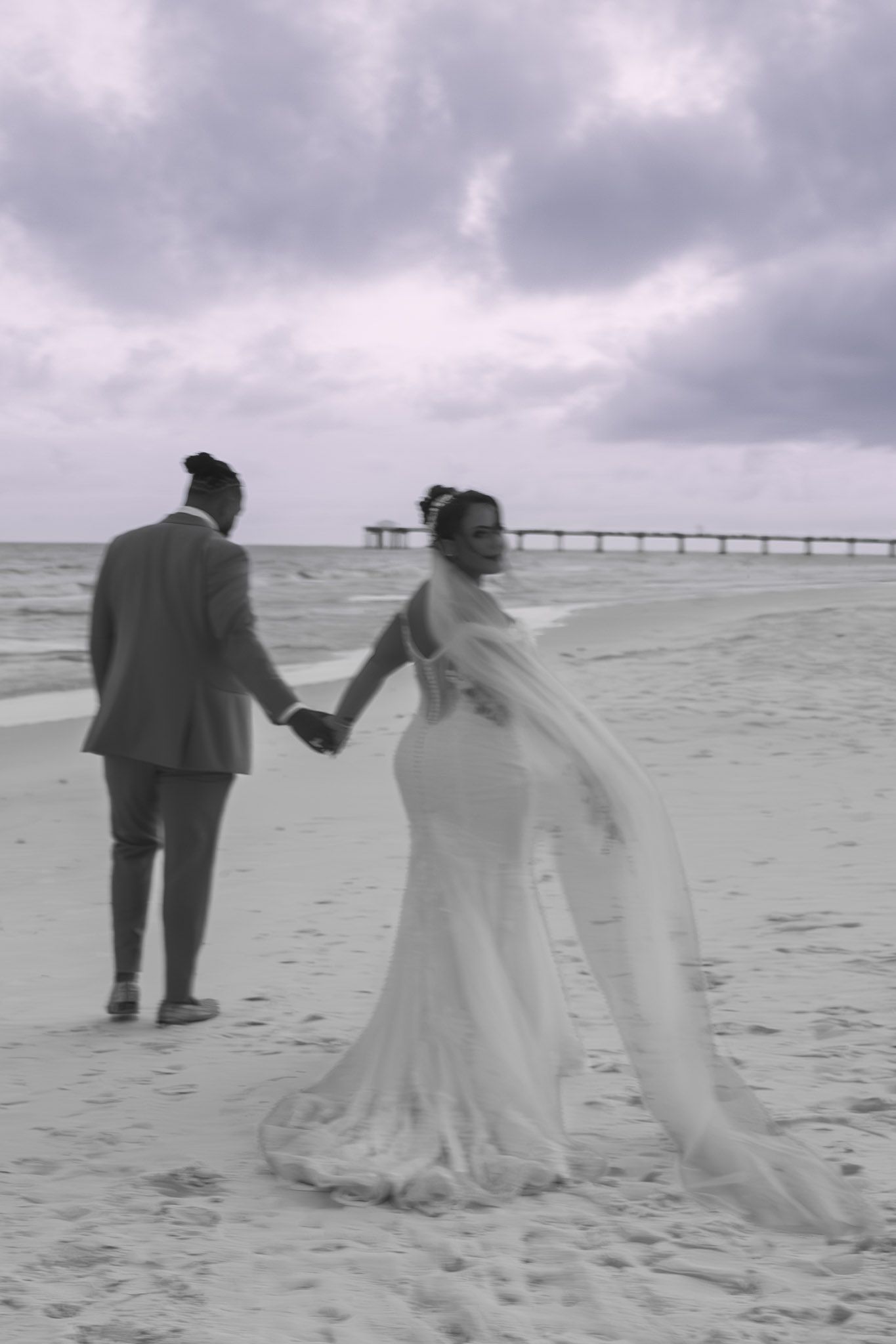 Black and white photo of a bride and groom holding hands on a beach, with a pier and cloudy sky in the background.