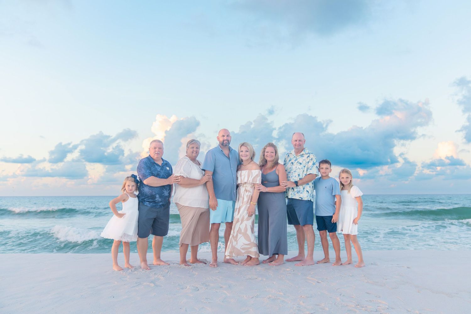 Family of nine poses on a sandy beach at sunset with calm ocean waves and pastel sky in the background.