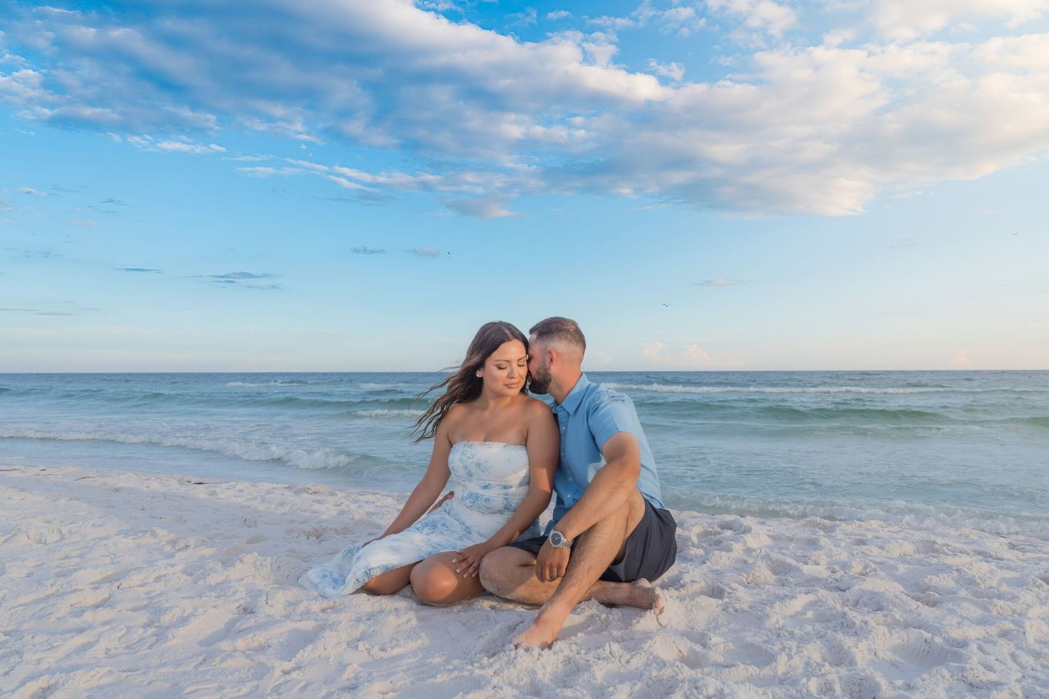 Couple sitting together on white sandy beach at sunset, waves and blue sky in background.