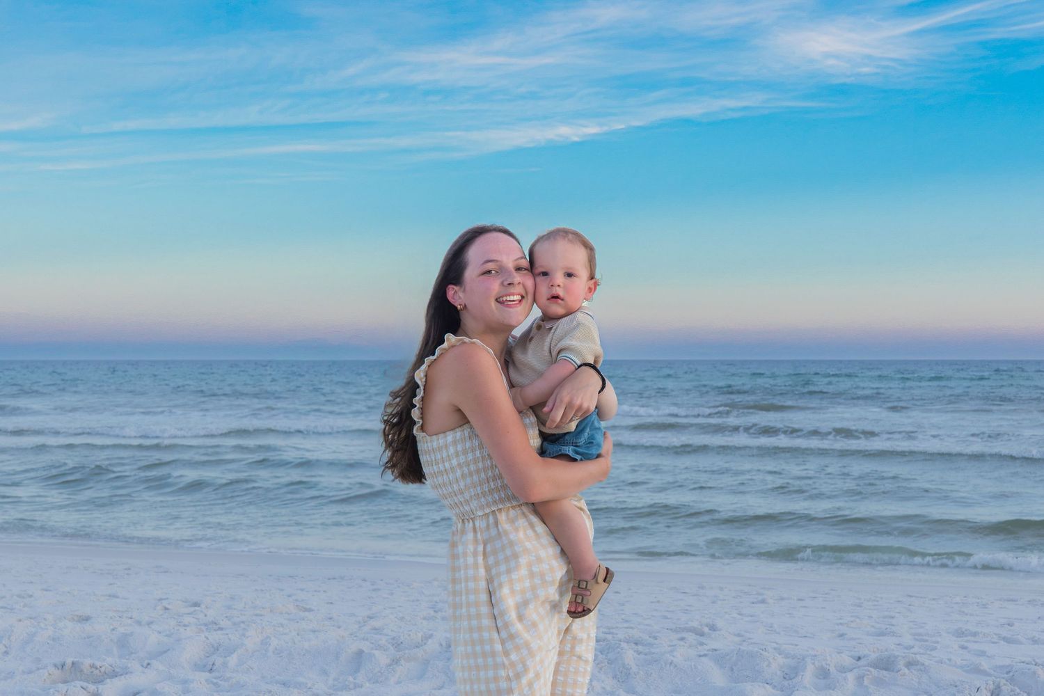 A young woman in a white dress holds a smiling baby on a sunny beach with calm ocean waves and a blue sky.